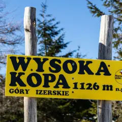 Żółta tablica na dwóch drewnianych słupkach informuje: "Wysoka Kopa 1126 m n.p.m. Góry Izerskie".