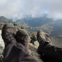 Stopy w butach trekkingowych na tle jesiennych Tatr. Październikowe słońce przebija się przez chmury, oświetlając skaliste szczyty i turkusowe jezioro.