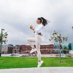 Kobieta w białym stroju sportowym wykonuje ćwiczenie, podnosząc kolana. W tle miejski krajobraz z budynkami i zielenią. To jeden z rodzajów skipów.