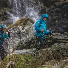 Dwie osoby w turkusowych kurtkach przeciwdeszczowych i z kijami trekkingowymi idą po kamienistym szlaku przy wodospadzie.