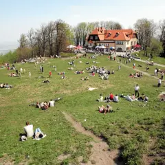 Ludzie odpoczywają na trawie na zboczu góry. W tle budynek z czerwonym dachem. Widać ścieżki i drzewa.