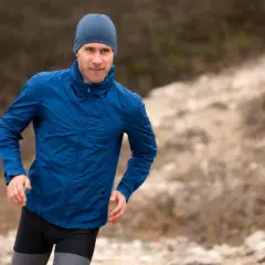 Mężczyzna w niebieskiej kurtce softshell i czapce biegnie po kamienistym terenie. Pamiętaj, jak prać softshell, by służył Ci latami.