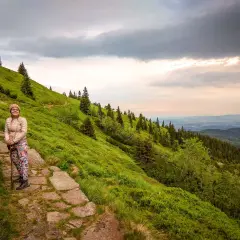 Kobieta na kamiennym szlaku, podziwiająca widok z zielonego zbocza. W oddali widać lasy i góry, a nad nimi burzowe chmury. To piękny widok ze **śnieżka szczyt**.