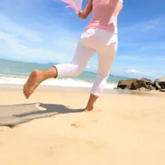 Kobieta w białych spodniach i różowej bluzce cieszy się bieganiem boso po piaszczystej plaży, z falującym morzem i błękitnym niebem w tle.