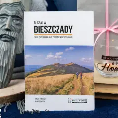 Przewodnik "Ruszaj w Bieszczady" z widokiem na góry i turystów. Obok rzeźba z drewna i świeca z napisem "all you need is in your Home".