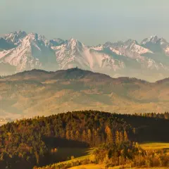 Widok na ośnieżone szczyty górskie, przed nimi łagodne wzgórza pokryte jesiennymi lasami. Lubań góra w oddali.