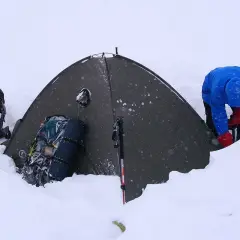 Osoba w niebieskiej kurtce i czerwonych rękawiczkach przygotowuje się do biwaku w śniegu. Obok namiotu stoją plecaki i kije trekkingowe.