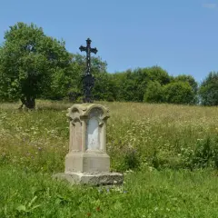 Kamienny postument z krzyżem na wzgórzu porośniętym dzikimi kwiatami. W tle las i błękitne niebo. Dźwiniacz górny.