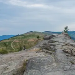 Kamienista ścieżka prowadzi na beskid slaski szczyt. W oddali widać zielone wzgórza i niebieskie pasma górskie pod zachmurzonym niebem.