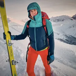 Anna figura w narciarskim stroju, z nartami w ręku, na tle ośnieżonych gór.