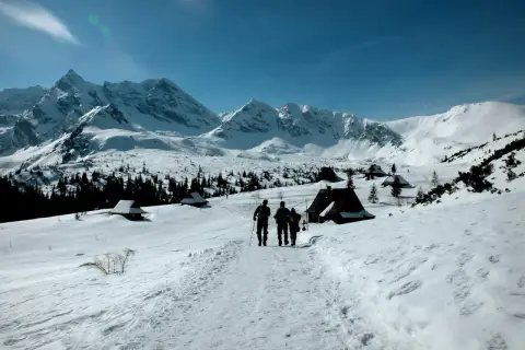 Trzej turyści idą szlakiem przez zasypane śniegiem Tatry zimą dla średniozaawansowanych. W oddali widać ośnieżone szczyty i górskie chatki.