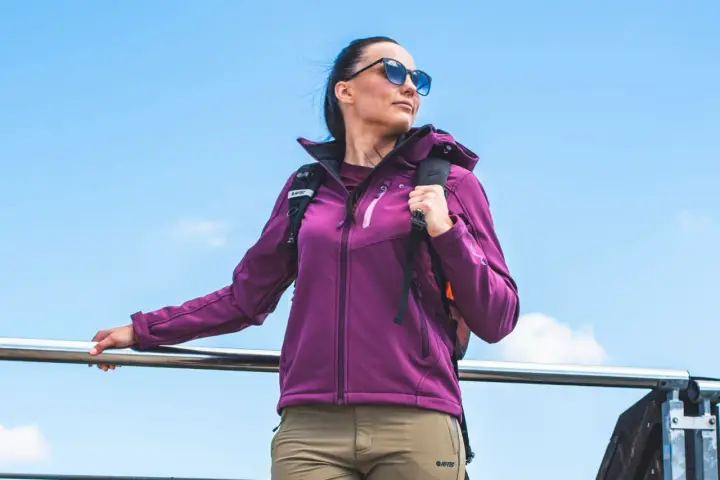Kobieta w fioletowej kurtce softshell i beżowych spodniach, z plecakiem, na tle błękitnego nieba. Idealna na wędrówkę, lepsza niż hardshell.