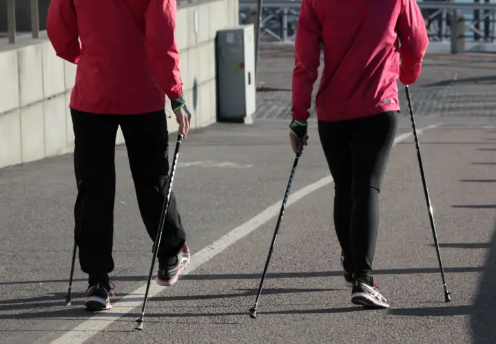 Dwie osoby w czerwonych kurtkach i czarnych spodniach idą z kijkami do nordic walking. Pokazują, jak ustawić kijki do chodzenia.