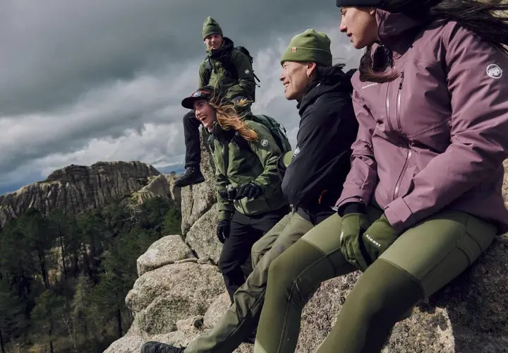 Grupa przyjaciół w zielonych i fioletowych kurtkach softshell siedzi na skale, podziwiając górski krajobraz.