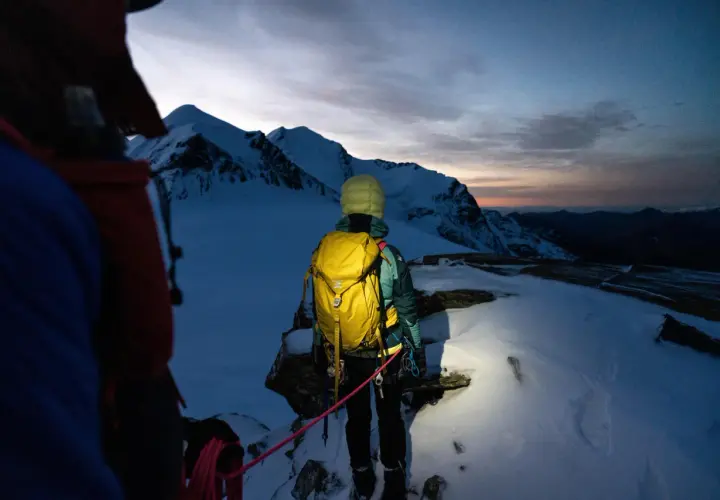 Alpinista z żółtym plecakiem lawinowym, gotowy na wyprawę w góry o wschodzie słońca. Plecak lawinowy jak działa? Zapewnia bezpieczeństwo w górach.