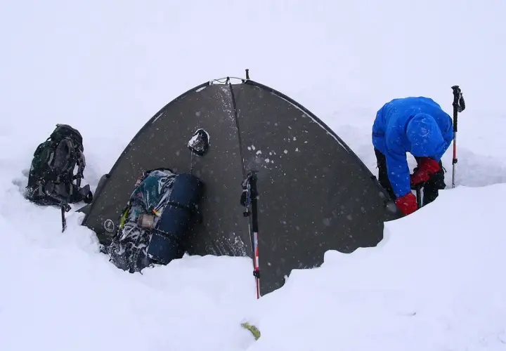 Osoba w niebieskiej kurtce i czerwonych rękawiczkach przygotowuje się do biwaku w śniegu. Obok namiotu stoją plecaki i kije trekkingowe.
