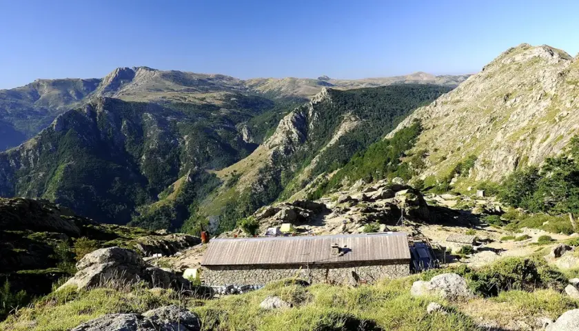 Kamienny schron na GR20 Korsyka, otoczony skalistymi zboczami i zielonymi dolinami.