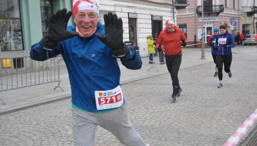 Uśmiechnięty biegacz z numerem 571 na piersi, macha rękami. W tle inni uczestnicy biegu, tworzący żywy kalendarz biegów górskich.