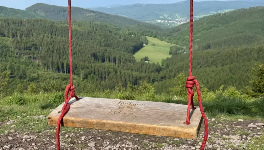 Drewniana huśtawka z napisem "Ruprechticki szpiczak" wisi nad malowniczą doliną pełną zielonych lasów i pól.