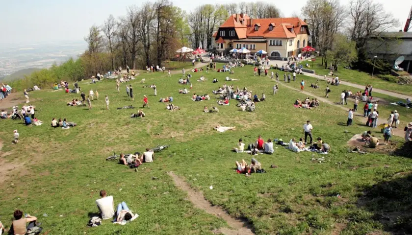 Ludzie odpoczywają na trawie na zboczu góry. W tle budynek z czerwonym dachem. Widać ścieżki i drzewa.