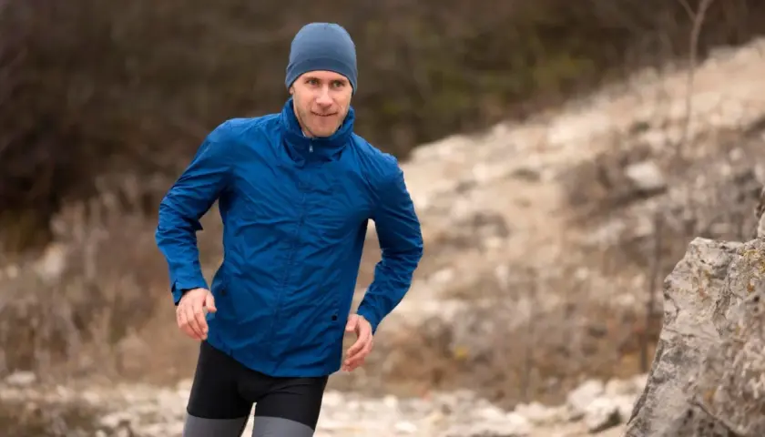 Mężczyzna w niebieskiej kurtce softshell i czapce biegnie po kamienistym terenie. Pamiętaj, jak prać softshell, by służył Ci latami.