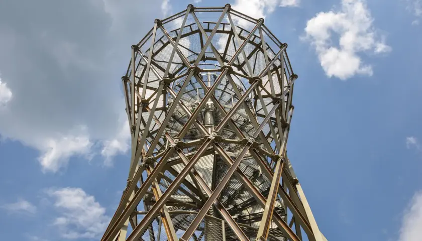 Metalowa konstrukcja wieży widokowej, przypominająca spiralę, wznosi się ku niebu. W oddali widać borowa góra.