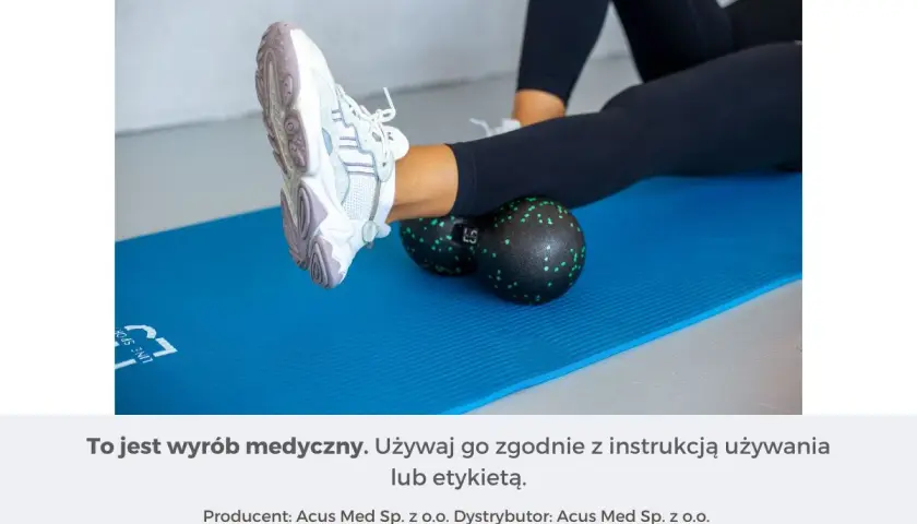 Kobieta wykonuje rolowanie mięśni łydki za pomocą czarnego rollera z zielonymi kropkami, leżąc na niebieskiej macie.