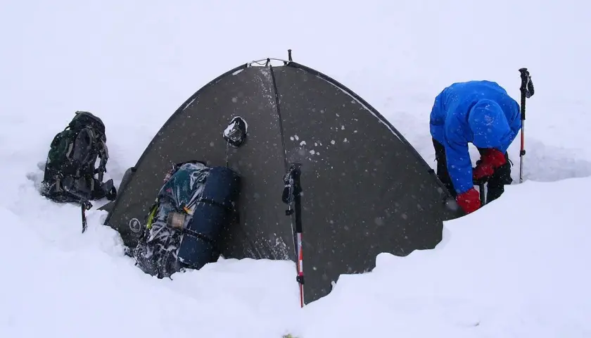 Osoba w niebieskiej kurtce i czerwonych rękawiczkach przygotowuje się do biwaku w śniegu. Obok namiotu stoją plecaki i kije trekkingowe.