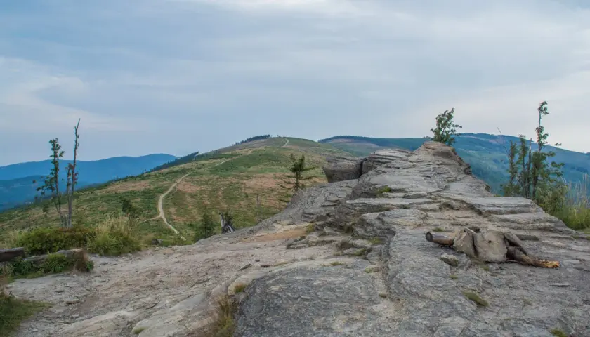 Kamienista ścieżka prowadzi na beskid slaski szczyt. W oddali widać zielone wzgórza i niebieskie pasma górskie pod zachmurzonym niebem.