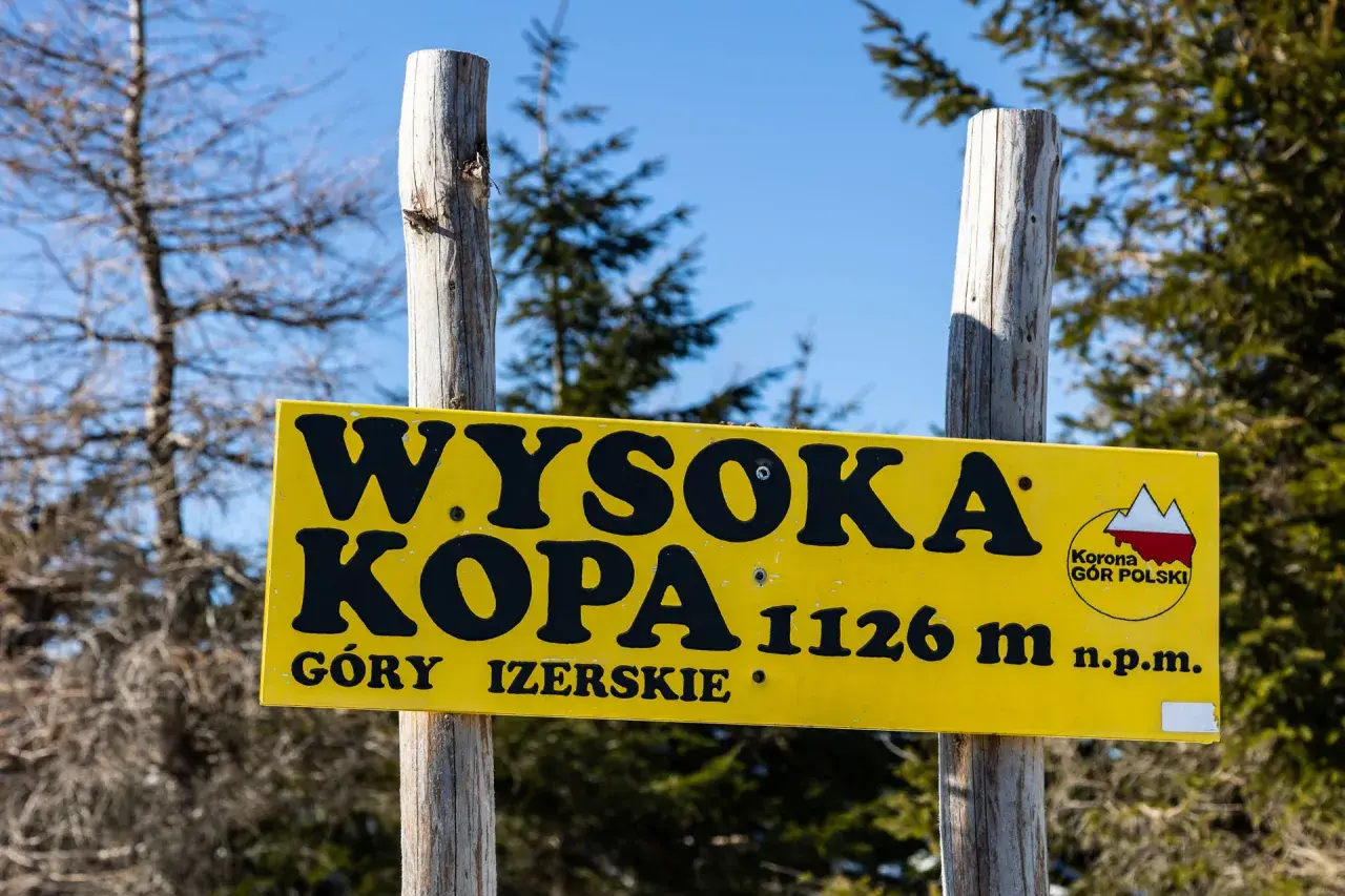 Żółta tablica na dwóch drewnianych słupkach informuje: "Wysoka Kopa 1126 m n.p.m. Góry Izerskie".