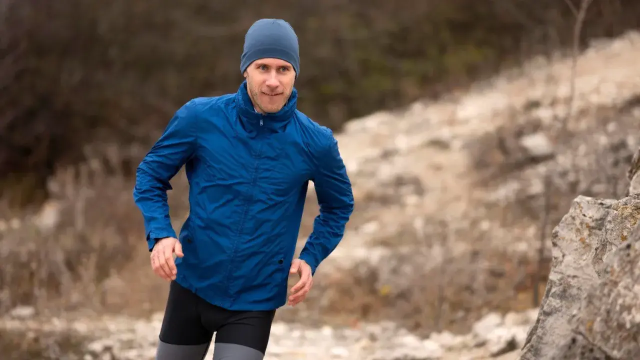 Mężczyzna w niebieskiej kurtce softshell i czapce biegnie po kamienistym terenie. Pamiętaj, jak prać softshell, by służył Ci latami.