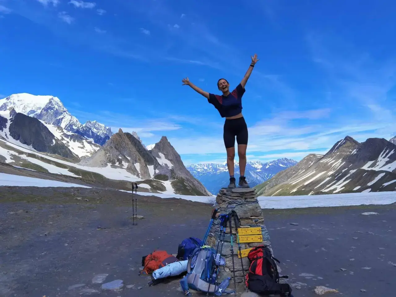 Szczęśliwa turystka na szczycie kamiennego kopca, z plecakami i kijkami trekkingowymi u stóp, na tle majestatycznych Alp podczas ultra-trail du mont-blanc.