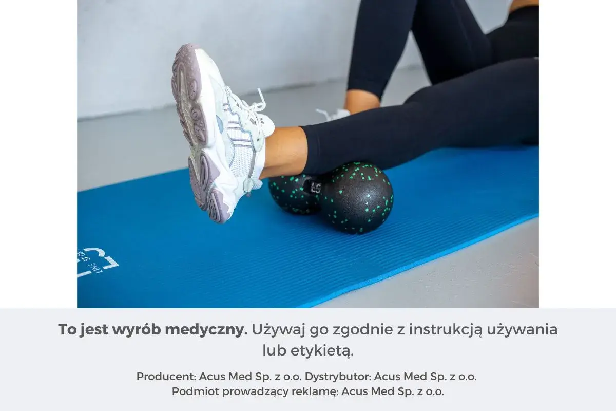 Kobieta wykonuje rolowanie mięśni łydki za pomocą czarnego rollera z zielonymi kropkami, leżąc na niebieskiej macie.