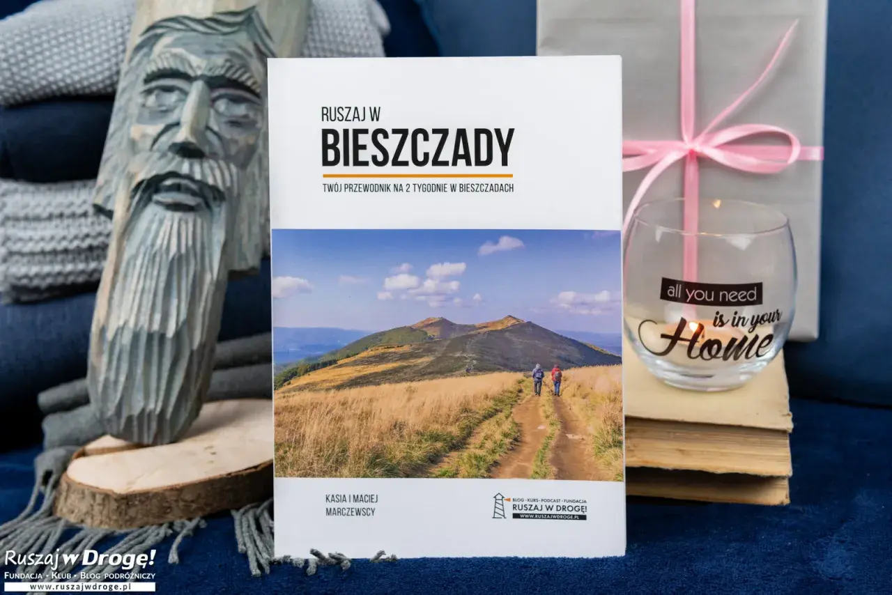 Przewodnik "Ruszaj w Bieszczady" z widokiem na góry i turystów. Obok rzeźba z drewna i świeca z napisem "all you need is in your Home".