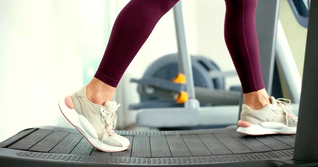Kobieta w bordowych legginsach i beżowych butach ćwiczy na bieżni. Czy bieżnia odchudza brzuch? Regularny trening cardio, jak bieganie, wspiera redukcję tkanki tłuszczowej.