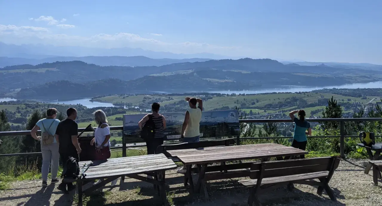 Widok na Jezioro Czorsztyńskie i góry. Grupa turystów podziwia panoramę, a tablica informacyjna opisuje szczyty, w tym górę Wdżar.
